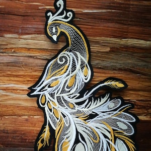 Op de afbeelding: Een geborduurde pauwpatch in wit en goud met ingewikkelde details. De pauw kijkt naar links en heeft zijn staartveren uitgespreid. De patch is op een bruine houten achtergrond.