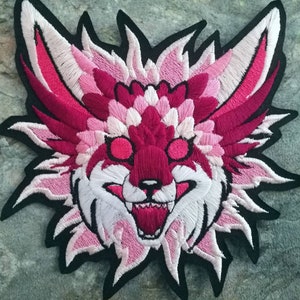 Peut inclure: Un écusson brodé représentant une tête de créature stylisée avec des oreilles pointues, des dents acérées et une gueule ouverte. Le motif utilise une palette de couleurs vives magenta profond, rose vif, rose pâle et blanc, bordé de noir.