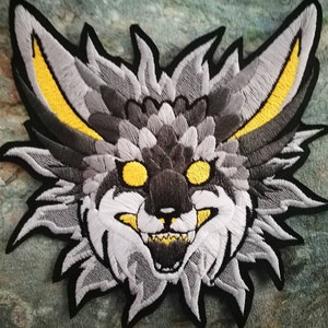 May include: Embroidered patch of a grey and black furry creature with yellow ears and glowing yellow eyes. The creature has a wide open mouth with sharp teeth.