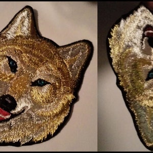 May include: Two embroidered patches of a wolf's head. The wolf has a light brown and white fur pattern with blue eyes. One patch is facing forward and the other is facing sideways.