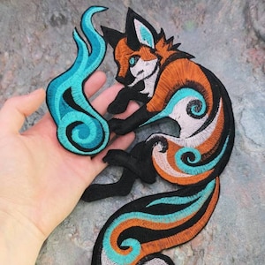 Puede incluir: Parche bordado para planchar de un zorro estilizado naranja y blanco con una cola de llama azul y turquesa. El zorro está sentado sobre sus cuartos traseros con la cola enroscada alrededor de su cuerpo.