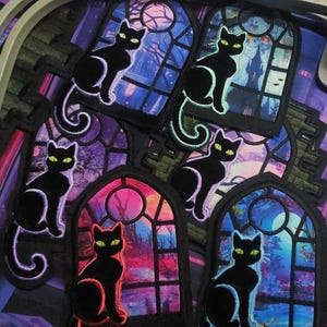 May include: Embroidered patches featuring black cats with glowing green eyes, perched in front of colorful stained-glass window scenes. The windows depict night scenes with moons, castles, and trees. The patches have a gothic or Halloween theme.