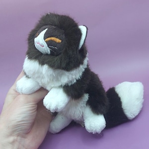 May include: A plush cat toy featuring long, soft fur in dark brown and white. The cat has a white muzzle, a small pink embroidered nose, and closed eyes with orange stitching. It has a bushy tail with alternating dark brown and white sections.