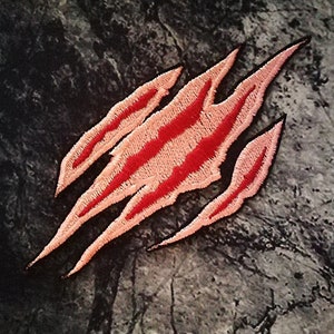 May include: Pink and red embroidered claw mark patch. The patch is shaped like a claw with three points and has a red outline. The claw is on a grey background.