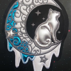 Op de afbeelding: Een zilveren kat die op een witte halve maan zit met blauwe en zilveren swirls. De maan druppelt met witte en blauwe vloeistof. Er zijn zilveren sterren op de achtergrond.