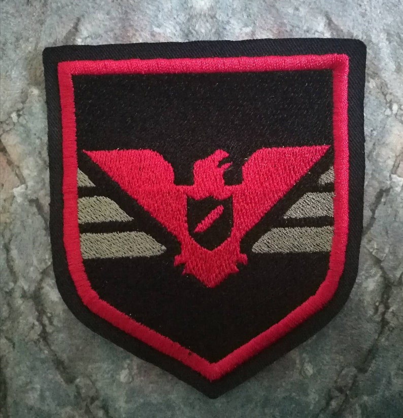 May include: Embroidered patch with a shield shape, featuring a red eagle design on a black background. The patch has a red border and a gray horizontal stripe. The patch is likely for a jacket or bag.