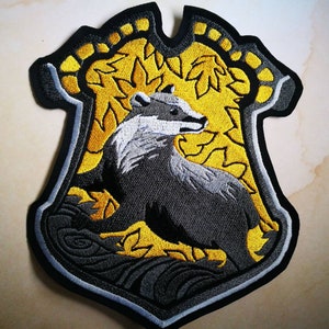 Puede incluir: Un parche bordado negro y gris con un tejón sobre un fondo amarillo y dorado. El parche tiene un contorno blanco y una forma de escudo.