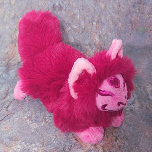 Puede incluir: Un zorro de peluche rosa y rojo con un dise&ntilde;o en forma de coraz&oacute;n en la cara. El zorro est&aacute; acostado de lado con la cabeza apoyada en las patas.