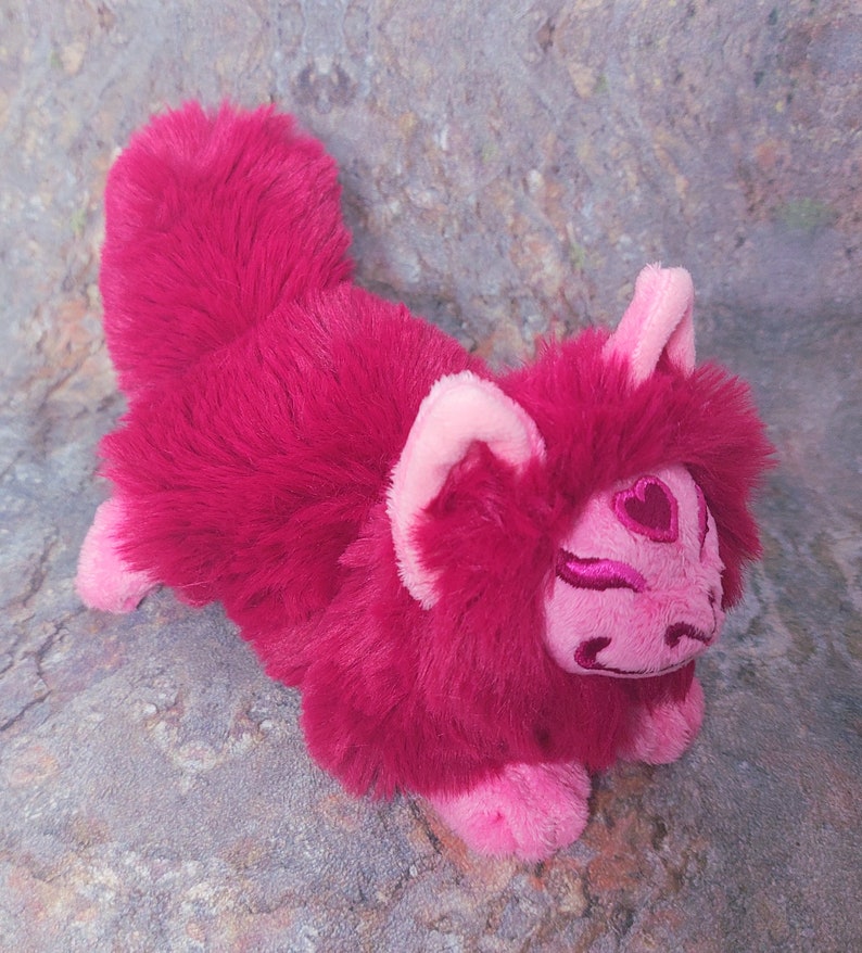 Puede incluir: Un zorro de peluche rosa y rojo con un dise&ntilde;o en forma de coraz&oacute;n en la cara. El zorro est&aacute; acostado de lado con la cabeza apoyada en las patas.