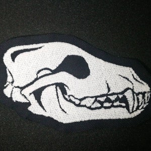 May include: White embroidered patch of an animal skull with teeth on a black fabric background.