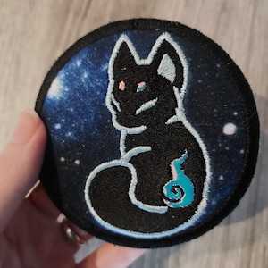 Op de afbeelding: Een zwarte geborduurde patch van een kat die op een blauwe en witte galaxy achtergrond zit. De kat heeft roze ogen en een blauwe en roze vlamstaart.