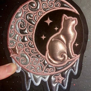 Op de afbeelding: Een roze en zilveren geborduurde patch van een kat die op een halve maan met sterren zit. De maan druppelt met zilver en roze.