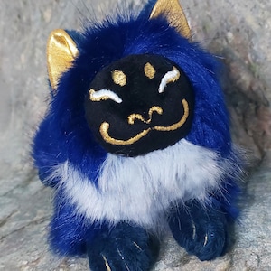 Puede incluir: Un gato de peluche azul y blanco con orejas doradas y una cara negra con bordados dorados. El gato est&aacute; acostado con las patas metidas debajo del cuerpo.