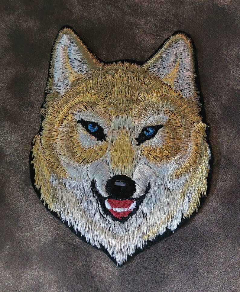 May include: Embroidered patch of a wolf's face with blue eyes and a red tongue. The wolf is depicted with a white and gold fur pattern.