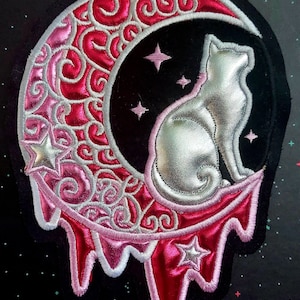 May include: A pink and white embroidered patch of a silver cat sitting on a dripping crescent moon with stars.
