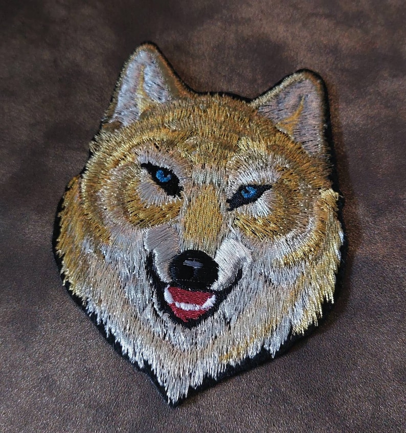 May include: Embroidered patch of a wolf's face with a white and gold fur pattern and blue eyes. The wolf is smiling with a red tongue.
