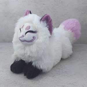 Puede incluir: Juguete de peluche de zorro blanco con orejas rosas y cola rosa. El zorro tiene una cara morada y blanca con un dise&ntilde;o estilizado.