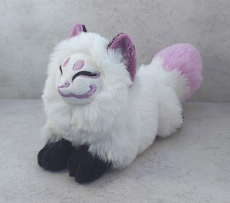 Puede incluir: Juguete de peluche de zorro blanco con orejas rosas y cola rosa. El zorro tiene una cara morada y blanca con un dise&ntilde;o estilizado.