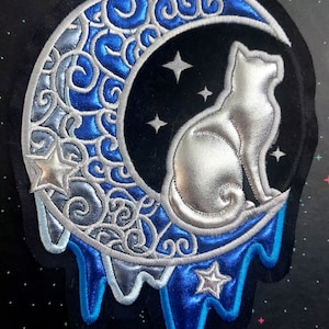 Op de afbeelding: Een zilveren kat die op een blauwe en zilveren halve maan zit met een druipende rand. De maan is versierd met een draaiend patroon en er zijn vijf witte sterren op de achtergrond.