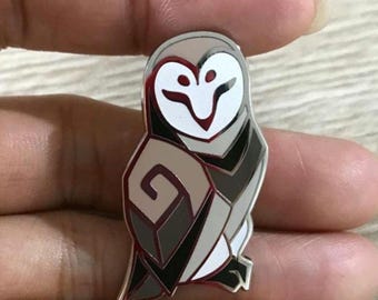 May include: A silver metal pin featuring a stylized owl with black and white geometric patterns. The owl has a white face with black eyes and a red beak.