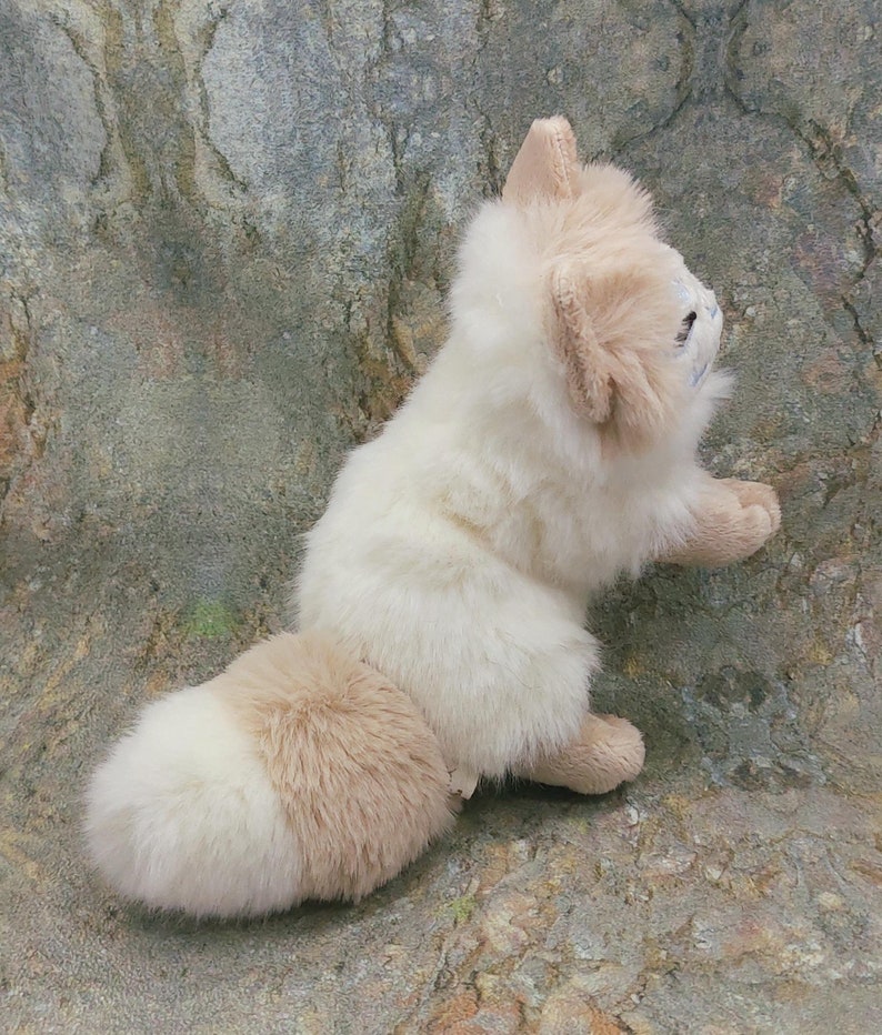 Puede incluir: Un zorro de peluche blanco y tostado con una cola esponjosa. El zorro est&aacute; sentado sobre una superficie gris texturizada.