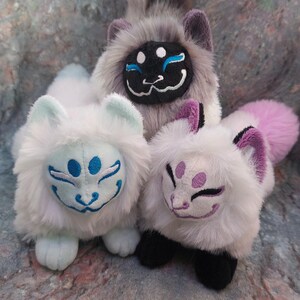 Puede incluir: Tres peluches con forma de zorro con marcas faciales de diferentes colores. El primer zorro es blanco con marcas azules, el segundo es gris con marcas negras y el tercero es blanco con marcas moradas.