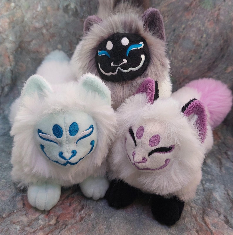 Puede incluir: Tres peluches con forma de zorro con marcas faciales de diferentes colores. El primer zorro es blanco con marcas azules, el segundo es gris con marcas negras y el tercero es blanco con marcas moradas.
