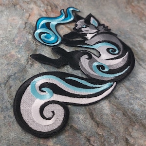 Puede incluir: Un parche bordado que representa un zorro estilizado en negro, blanco y azul. El zorro está enroscado con la cola envuelta alrededor de su cuerpo, y el diseño incluye patrones en espiral.