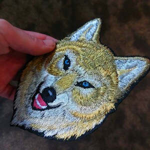 May include: Embroidered patch of a wolf's face with blue eyes and a white and gold fur pattern.