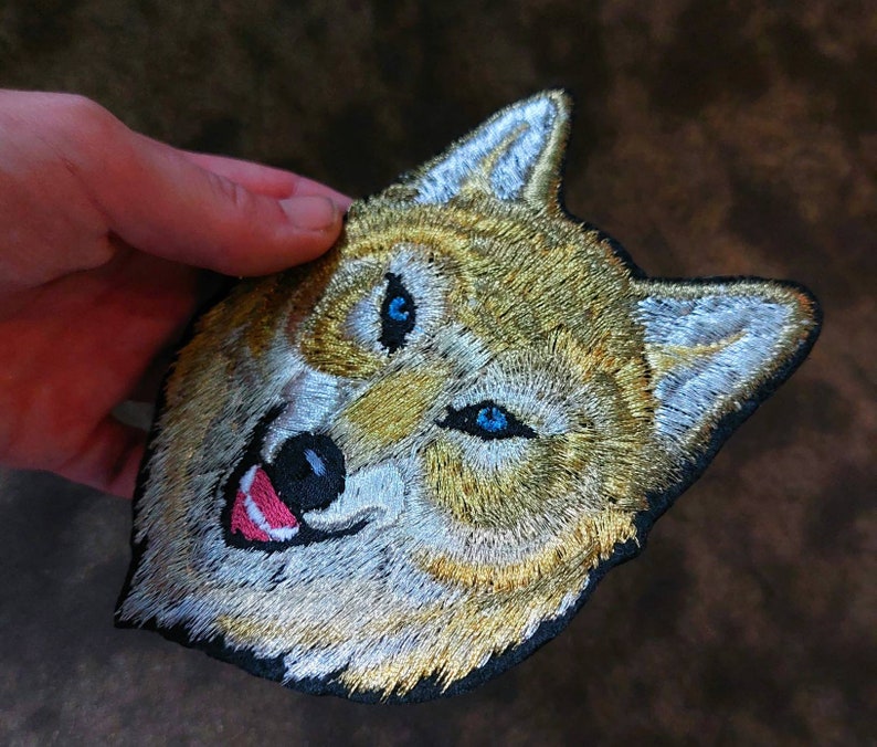 May include: Embroidered patch of a wolf's face with blue eyes and a white and gold fur pattern.