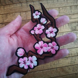 Könnte beinhalten: Gestickter Aufnäher mit einem Zweig rosa Kirschblüten mit weißen Akzenten. Der Aufnäher hat einen schwarzen Rand und braune Äste. Die Blüten befinden sich in verschiedenen Blühstadien und bilden ein detailliertes Blumendesign.