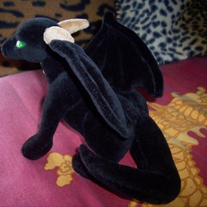 May include: Black plush dragon with green eyes and brown horns. The dragon is sitting on its hind legs with its wings spread out.