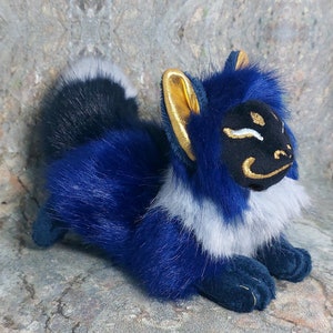 Puede incluir: Un zorro de peluche azul y blanco con orejas doradas y una m&aacute;scara negra en la cara. El zorro est&aacute; acostado de lado con la cabeza apoyada en sus patas.