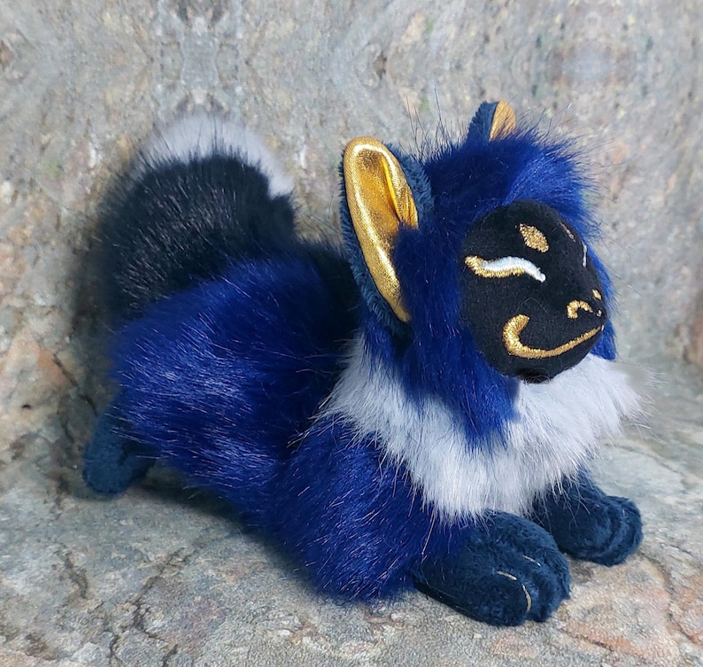 Puede incluir: Un zorro de peluche azul y blanco con orejas doradas y una m&aacute;scara negra en la cara. El zorro est&aacute; acostado de lado con la cabeza apoyada en sus patas.
