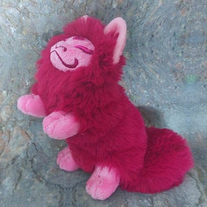 Puede incluir: Una criatura de peluche rosa y roja con forma de gato y una cola larga. El peluche tiene una cara sonriente con los ojos cerrados y una nariz rosa.