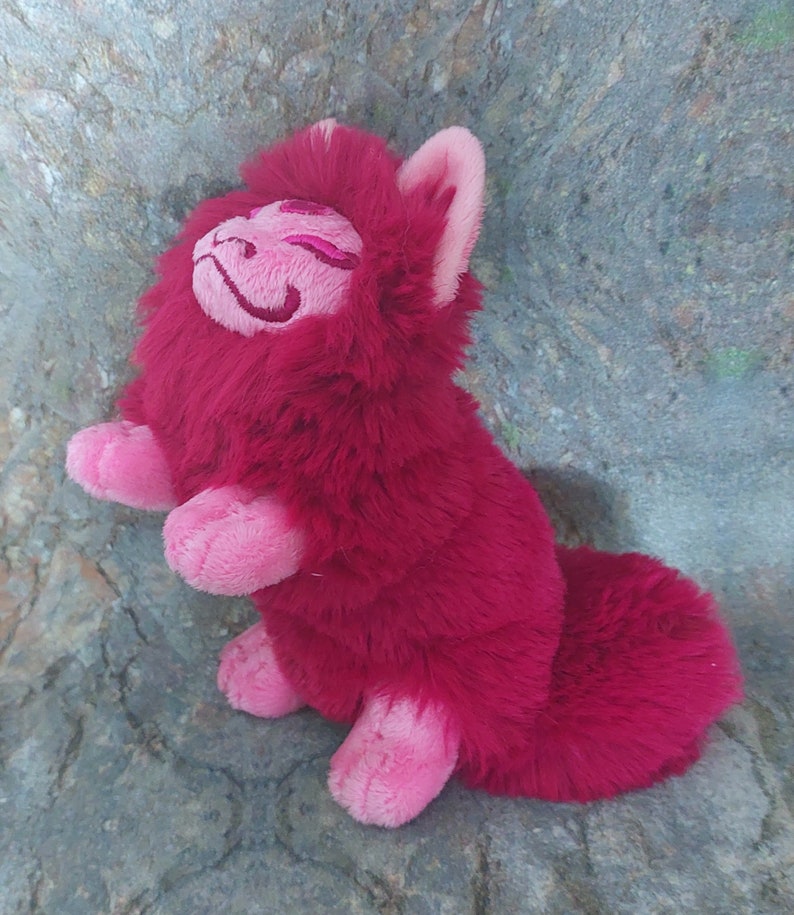 Puede incluir: Una criatura de peluche rosa y roja con forma de gato y una cola larga. El peluche tiene una cara sonriente con los ojos cerrados y una nariz rosa.