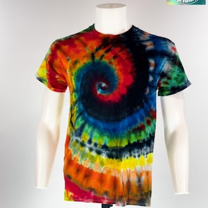 May include: A vibrant, tie-dye t-shirt in a spiral pattern, featuring a spectrum of colors including red, orange, yellow, green, blue, and black. The shirt is displayed on a mannequin. The logo "Cosmic Cool Dye Factory" is visible in the upper right corner.