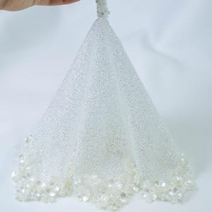 Puede incluir: Un velo de novia blanco y brillante con un ribete floral. El velo tiene forma de cono y está sujeto a un asa con cuentas. El velo está hecho de una tela transparente y brillante, perfecto para bodas y ocasiones especiales.