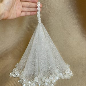 May include: A white conical veil featuring a fine glittery mesh fabric. The top of the veil is finished with a small loop of white floral embellishments, and the bottom edge is decorated with a dense border of white fabric flowers and beads.