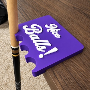 May include: A purple pool cue holder with white raised text that reads Nice Balls! The holder features three semi-circular cutouts along one edge to secure a pool cue, designed to rest on the edge of a flat surface.