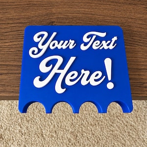 May include: A blue, trapezoidal, 3D-printed sign featuring the white, raised, cursive text "Your Text Here!" on the front. The bottom edge of the sign has four semi-circular cutouts, designed to hold items securely in place.