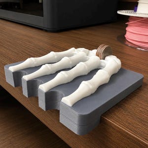 May include: A gray rectangular base with four white 3D-printed skeletal finger structures extending across the top surface. A single silver-colored coin is positioned upright between the two middle finger bones on the flat gray platform.