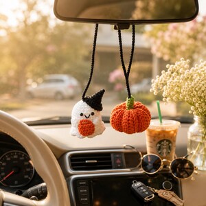 May include: Two crocheted hanging ornaments for a car rearview mirror. One is a white ghost wearing a black hat and holding a small orange pumpkin. The other is a round, textured orange pumpkin with a green stem. Both hang from black cords.