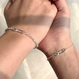 Könnte beinhalten: Zwei silberne Armbänder, jedes mit einem Sicherheitsnadel-Design. Eines hat eine Kettengliederkette, das andere eine Perlenkette. Die Armbänder werden an Handgelenken gezeigt. Ein stilvolles Accessoire.