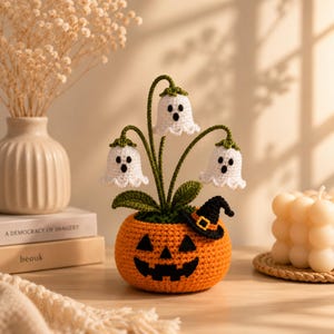 Könnte beinhalten: Ein gehäkelter orangefarbener Kürbis-Übertopf mit einem schwarzen Jack-o'-Lantern-Gesicht und einem kleinen schwarzen Hexenhut. Drei weiße gehäkelte Geisterblüten mit grünen Stielen und Blättern ragen oben heraus, jede mit schwarzen Augen und Mund.