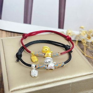 Könnte beinhalten: Zwei Armbänder mit Pferdeanhängern. Eines hat eine rote Kordel und einen goldenen Pferdeanhänger, das andere hat eine schwarze Kordel und einen silbernen Pferdeanhänger. Beide haben ein gewebtes blaues, orangefarbenes und gelbes Band.