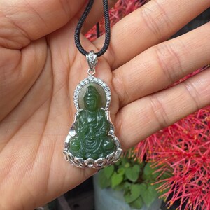 Op de afbeelding: Een donkergroene jade hanger gesneden in de vorm van een zittende figuur op een lotusbasis, gevat in een zilverkleurige metalen zetting met kleine heldere steentjes langs de boog, hangend aan een zwart koord met een zilverkleurige hanger.