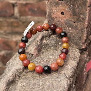Könnte beinhalten: Ein Armband mit Perlen aus verschiedenen farbigen Edelsteinen, darunter Rot, Braun, Gelb und Schwarz. Ein kleines Etikett mit dem Text "SL-710" ist angebracht. Das Armband wird auf einer rauen, strukturierten Oberfläche präsentiert.