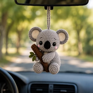 Könnte beinhalten: Ein graues, gehäkeltes Koala-Hängeornament mit schwarzer Nase, rosa Wangen und großen runden Ohren. Der Koala hält einen braunen, strukturierten Ast mit kleinen grünen Blättern. Es verfügt über eine gehäkelte Schlaufe zum Aufhängen im Fahrzeug.