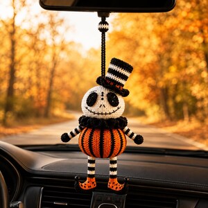 Könnte beinhalten: Ein gehäkelter Hängeschmuck mit einem weißen Totenkopf, schwarzen gestickten Augen und Mund sowie einem schwarz-weiß gestreiften Zylinder. Der Körper ist eine runde, orange-schwarze Kürbisform mit gestreiften Armen und geringelten Schuhen.